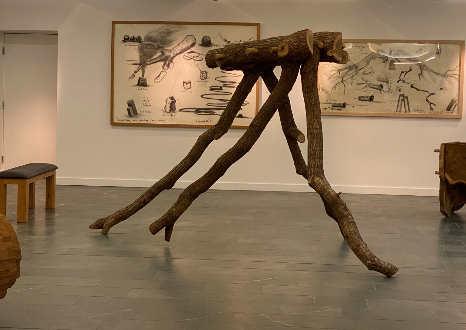The image shows a wooden sculpture resembling a tree trunk supported by four bowing legs in the centre of the frame, with drawings of tree trunks in frames behind it on the gallery wall.