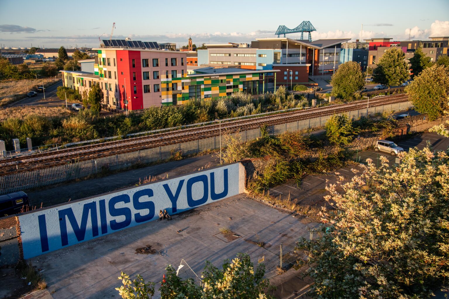A view of post-industrial Middlesbrough. In the foreground is a piece of wasteland with the words I miss you, painted in large blue letters against white, on a wall. Two figures are sitting close together against the wall. In the middle ground are railway tracks and some contemporary urban buildings. In the distance is the transporter bridge and a clear evening sky dotted with clouds. Warm light and long shadows indicate that the sun might set soon.