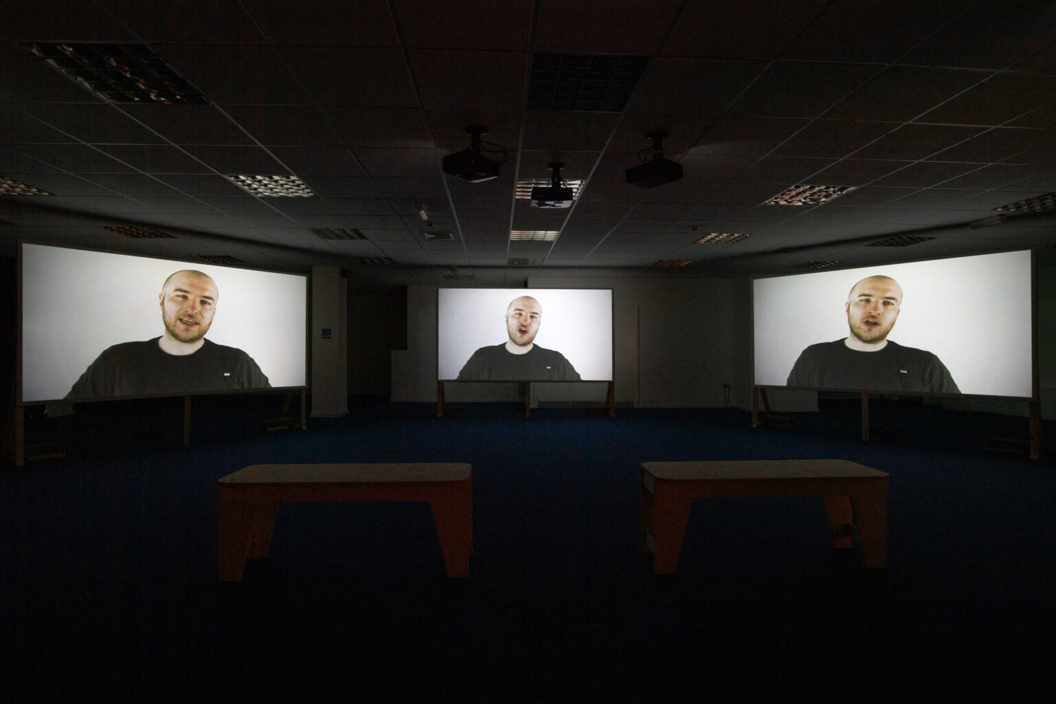 Three large screens in a dimly lit room. One on the far wall, in the middle of the photo, and the other two on adjacent walls either side. All three screens have a slightly similar image. A head and shoulders shot of a while male aged around 30. He has closely cropped hair, stubble and is wearing a black t-shirt. The background behind him is white. His face is different in each image as if he were talking or singing. There are two benches in the room in front of the screens.