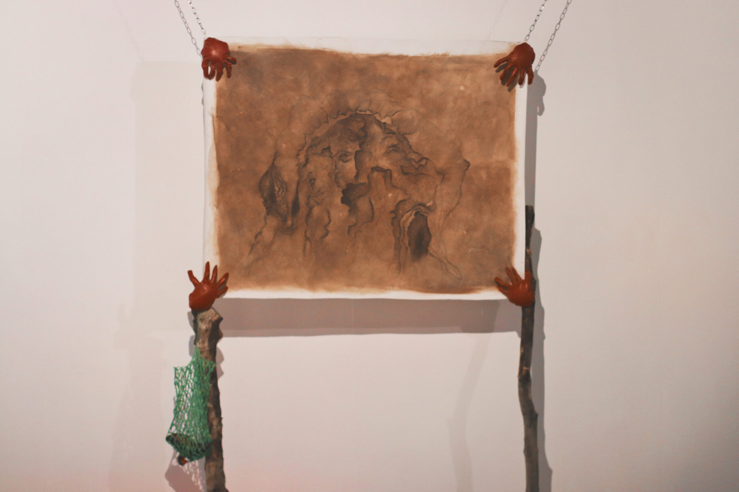 An image of a drawing on brown, distressed paper suspended by wires on top and gripped with hand-like clasps on the bottom.