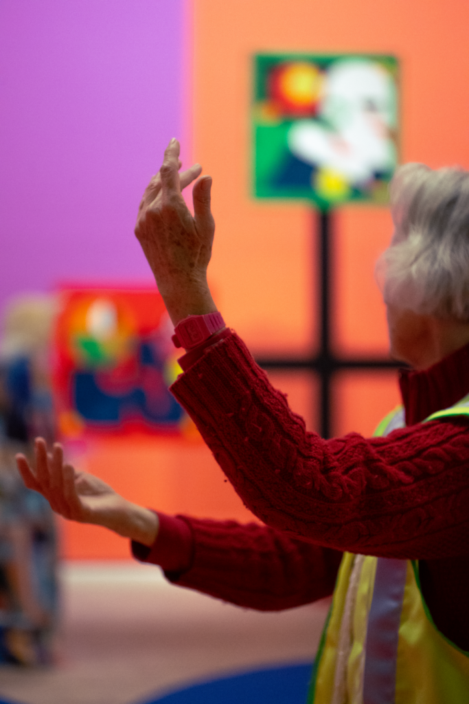 A woman with a silver bobbed hairstyle is facing away from the camera. She is wearing a red jumper, her arms are posed hands upwards. In the background are the colourful walls and furnishings of a bright installation.
