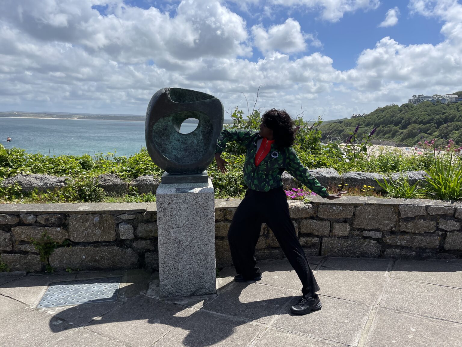 The artist stands beside a sculpture by Barbara Hepworth on a plinth, lifting her arm to touch it with her elbow; behind a coastal landscape on a sunny day..