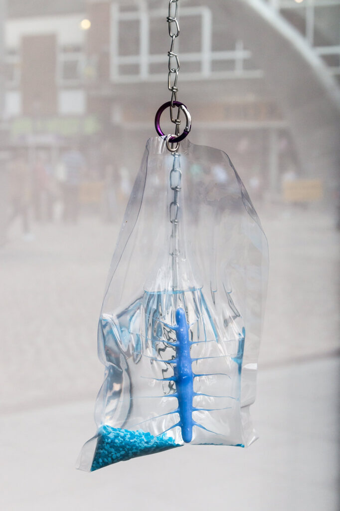 A vinyl bag filled with water, chlorine, and blue plastic shapes is suspended from above by a metal chain.