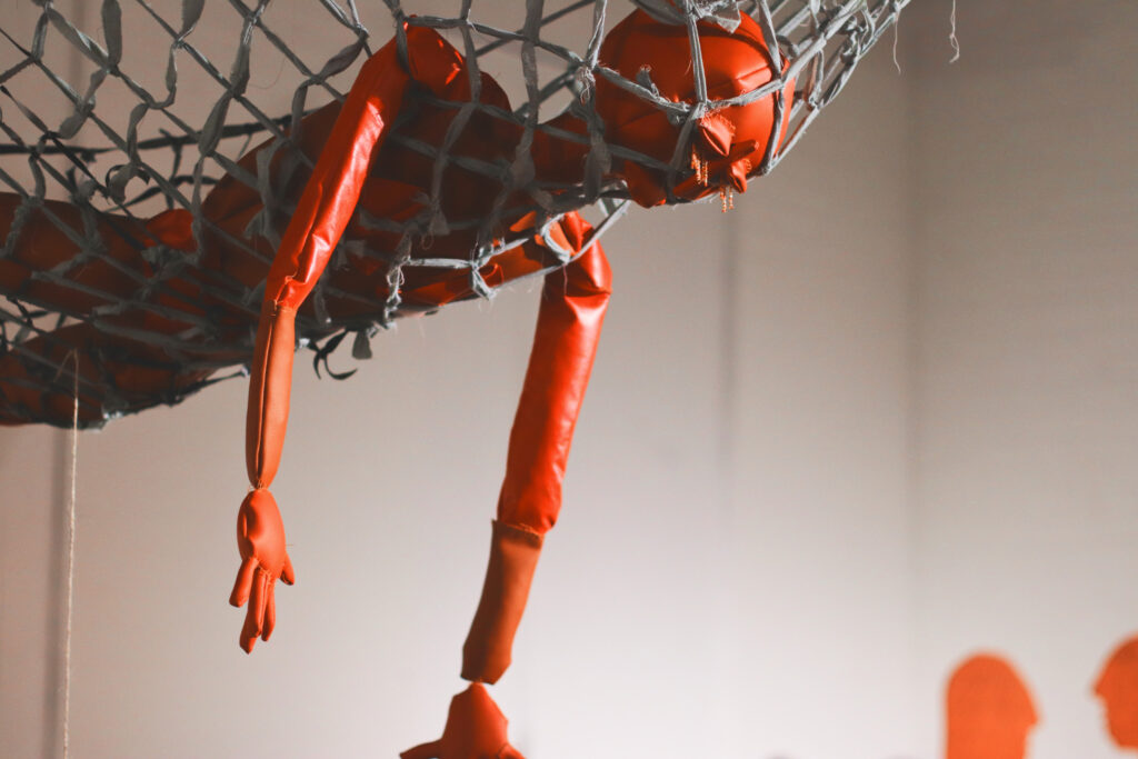 An image of a sculpture of a human form in red leather caught in a suspended net, with arms dangling towards the ground.