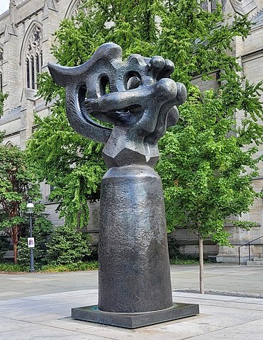 A bronze sculpture of abstract or semi-figural form in a leafy backdrop of a university campus.