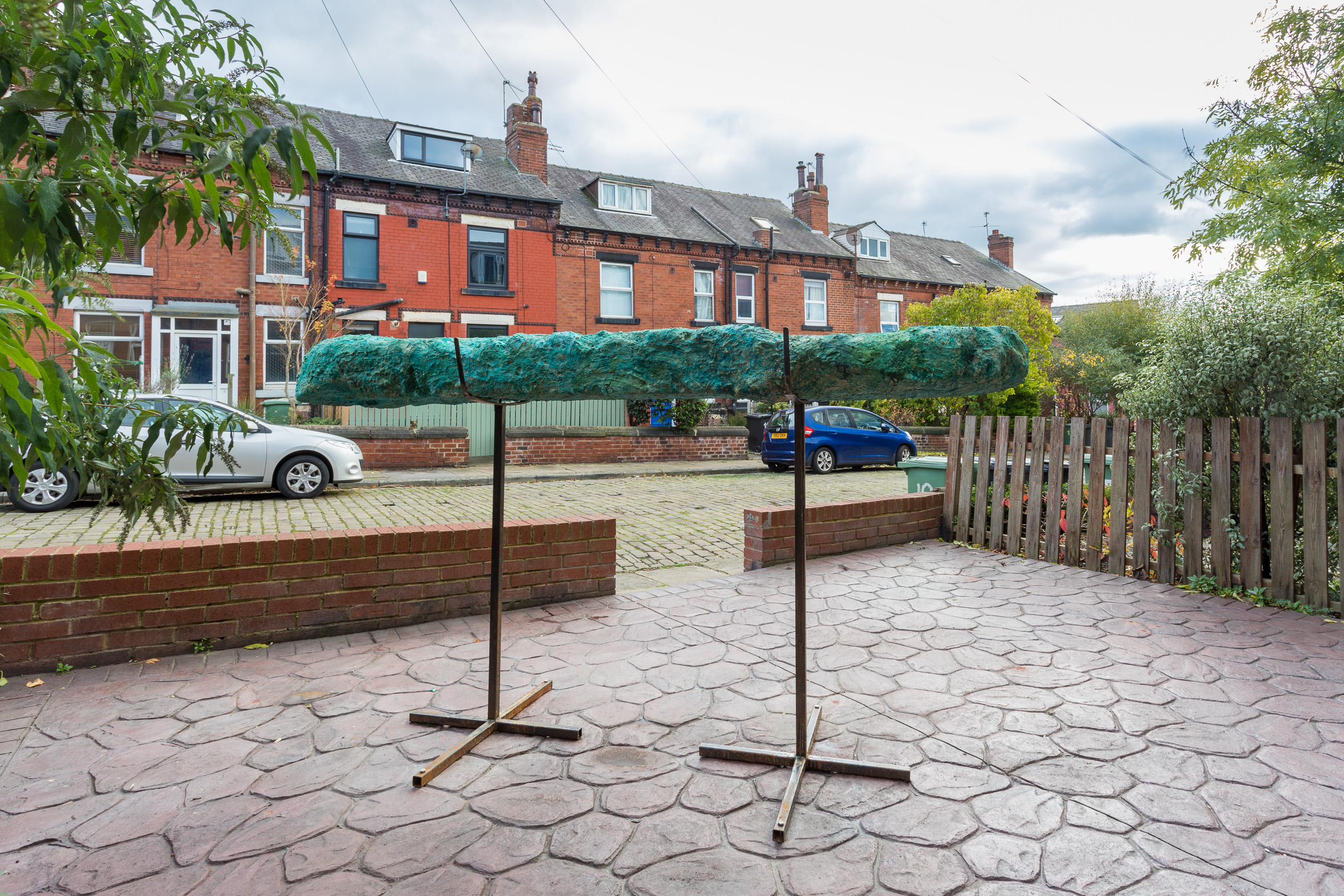 A beam like green sculpture is balanced on two metal supports in the paved back garden, with a view of a terraced back street.