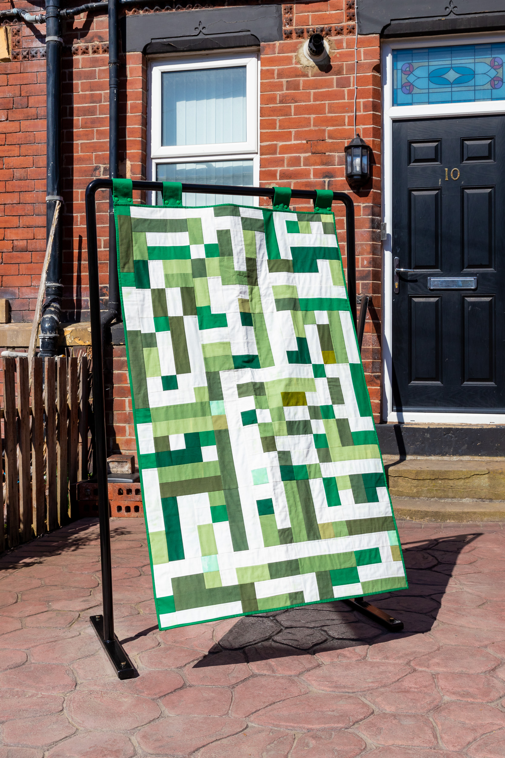 A geometric patterned textile is suspended in a black metal frame are in the paved back garden of a red-brick terraced house.