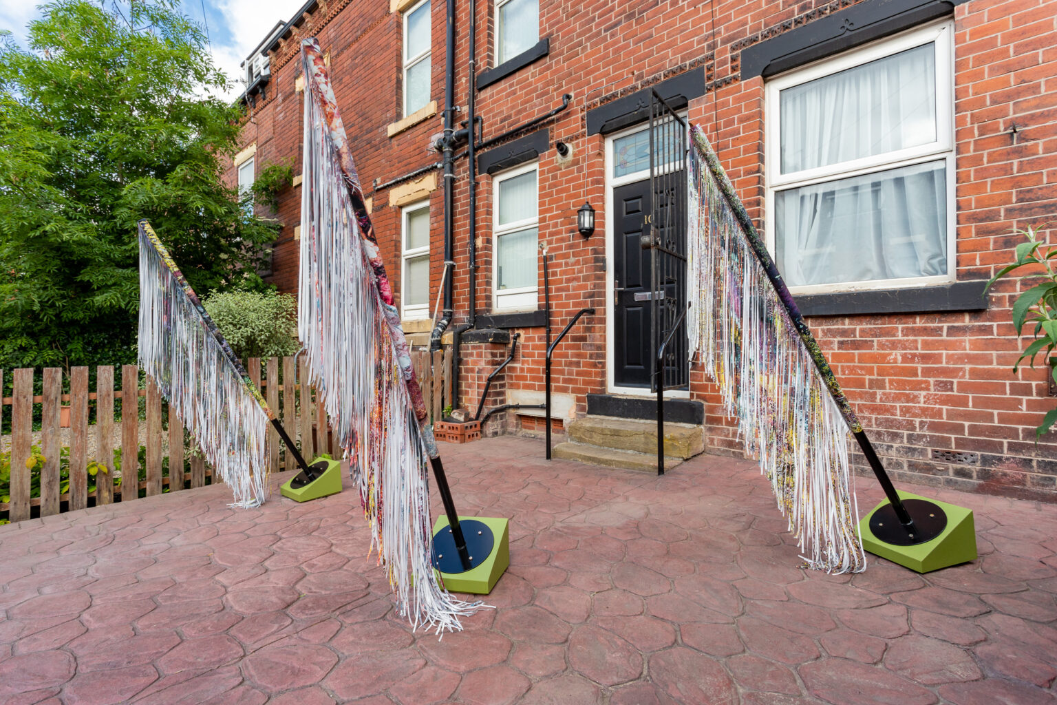 3 sculptures resembling flagpoles hung with fringed material are positioned in the paved back garden of a red-brick terraced house.