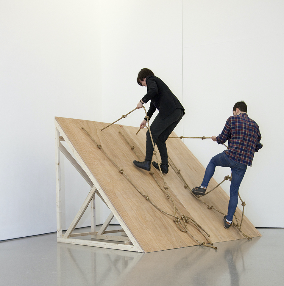 2 performers stand on a slanted board structure holding on with ropes in a white walled gallery space.