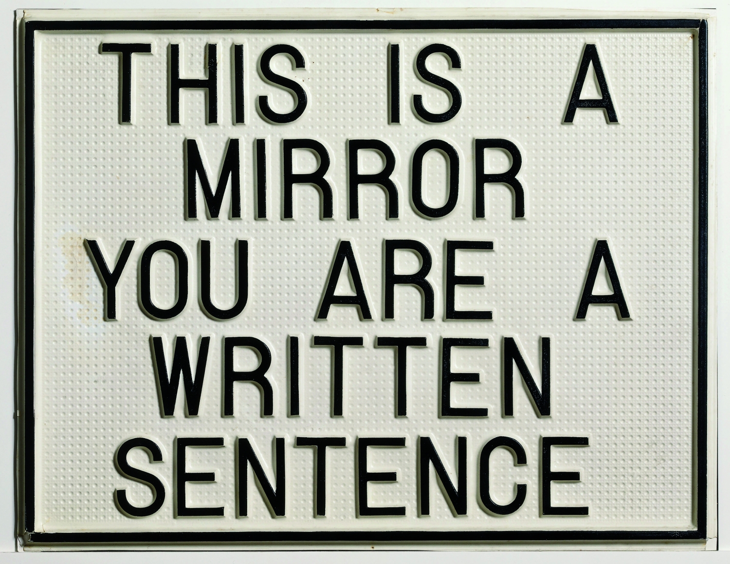 A small white plaque with a black border and caps letters spelling THIS IS A MIRROR YOU ARE A WRITTEN SENTENCE
