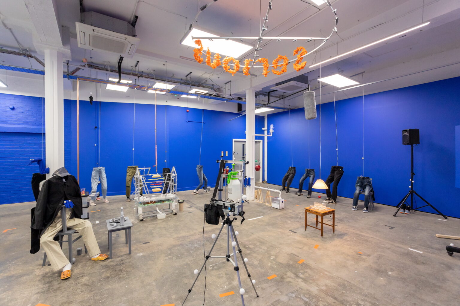 A view of a gallery space with blue walls and concrete floors and multiple artworks that resemble a film set