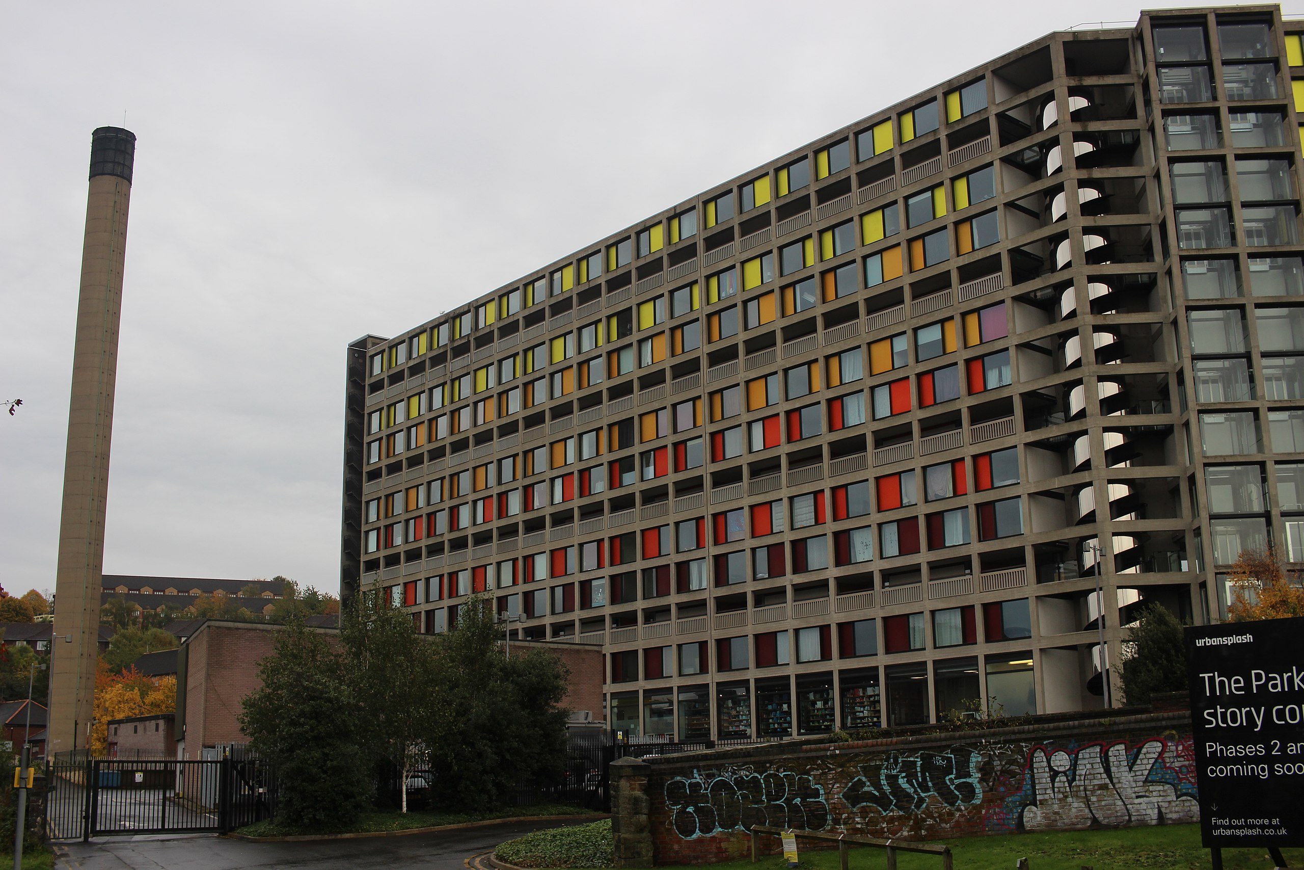 A concrete block of flats with rainbow cladding, against a grey sky and across from a smoke stack.