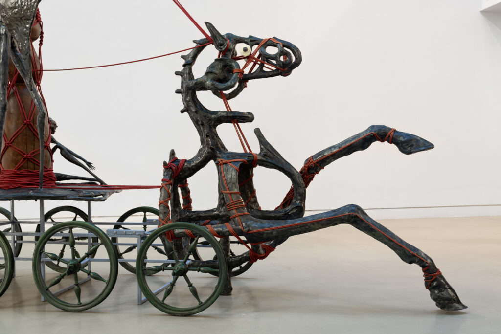 A skeletal iron sculpture of a horse pulling a cart with red twine pulling it backwards