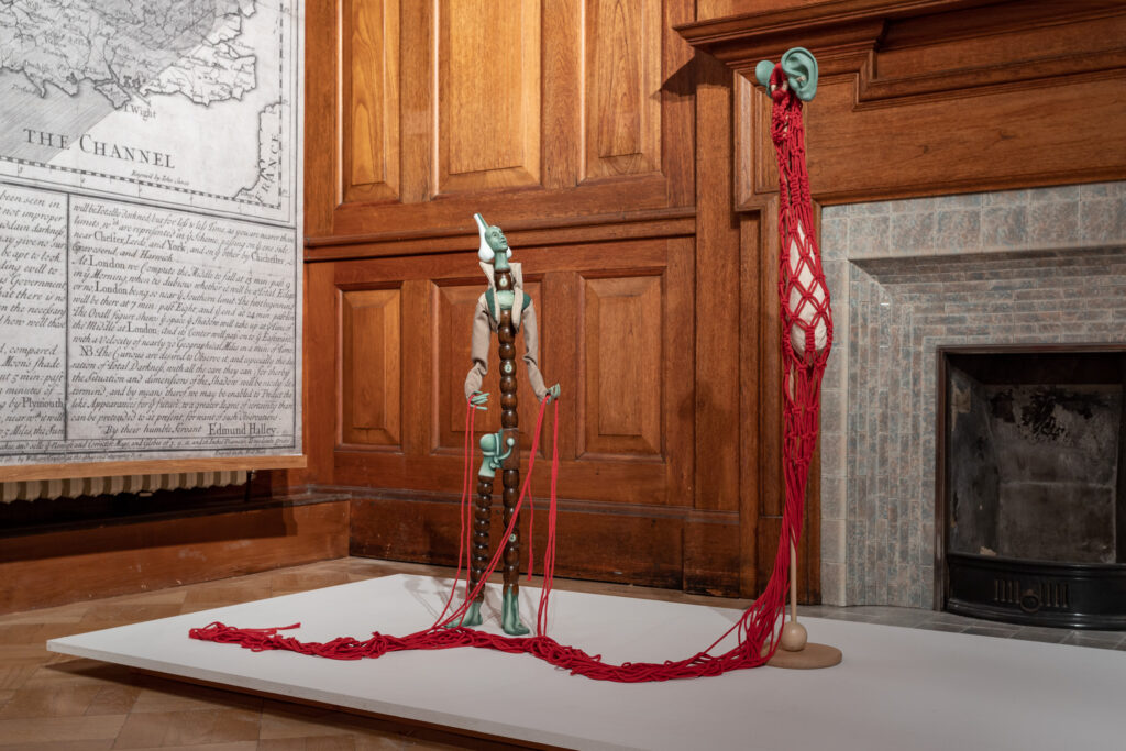 Thin tall sculptures sit in a wood panelled gallery beside an antique print hung on the wall