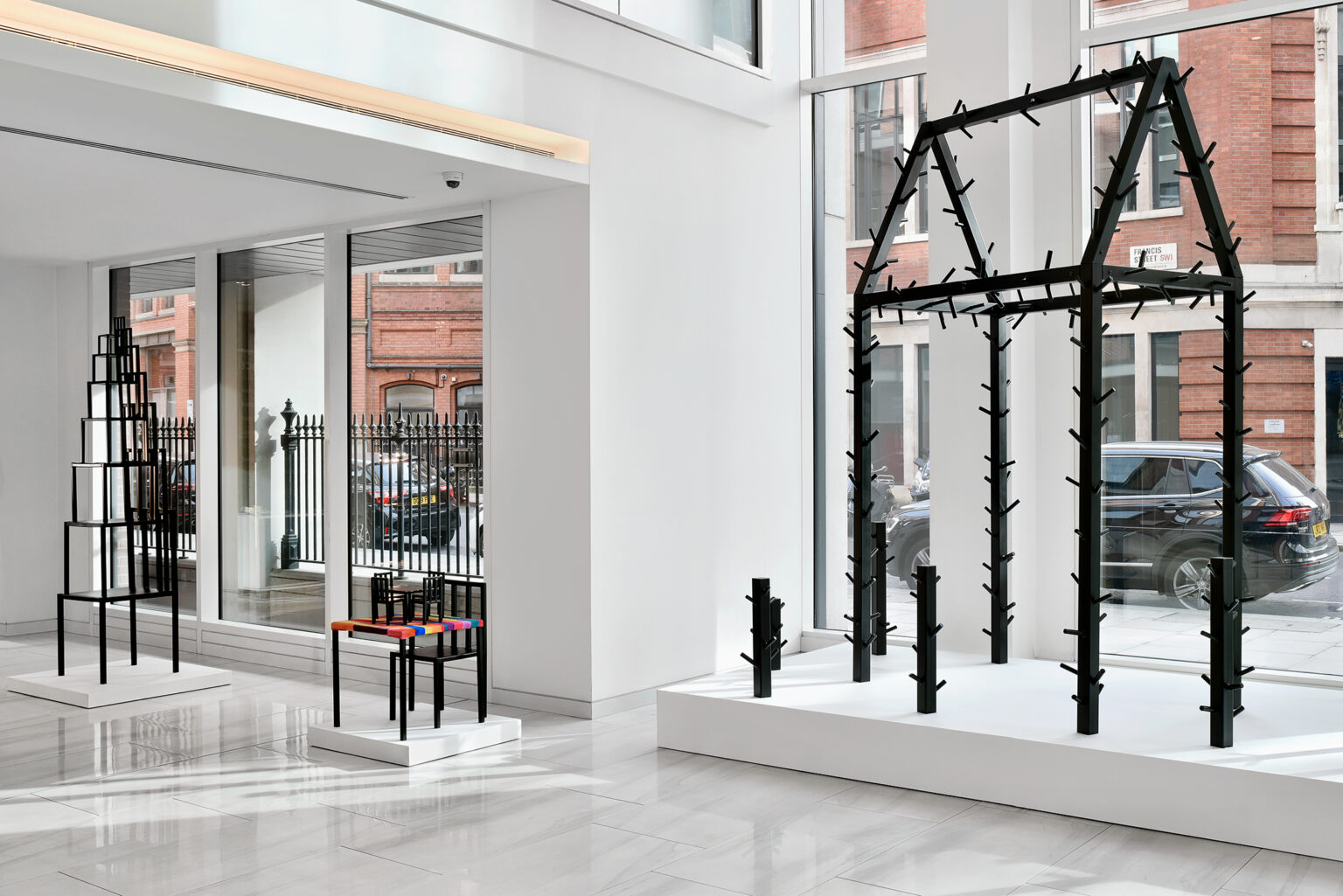 A windowed exhibition space with sculptures of black beams forming the outline of a house, with spikes on the outside of the beams