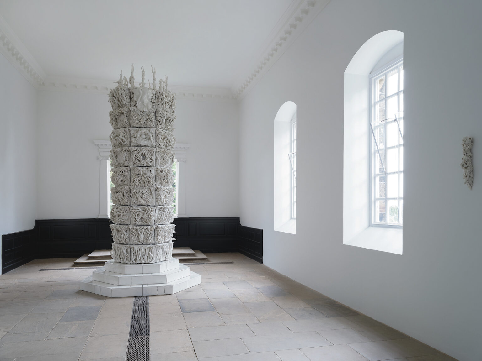A tall white column on a stepped plinth, covered in delicate sculptures, sits in the middle of a light gallery space with two windows.