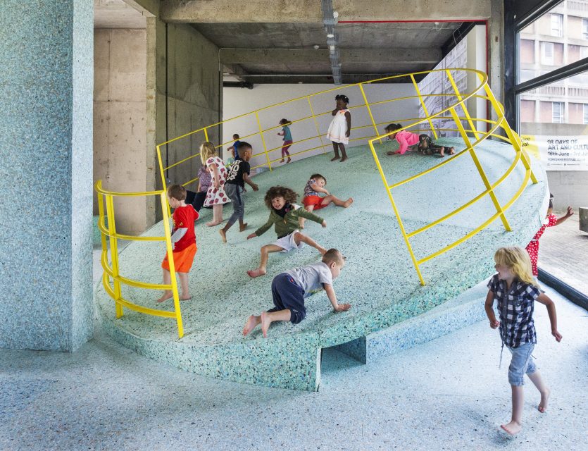 Children play on a large tilted foam circle with yellow railings in a concrete beamed exhibition space.