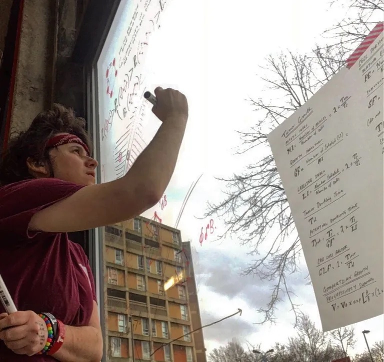 A person with short brown hair and a bandana and bracelets writes onto a window overlooking tower block flats and winter trees. 