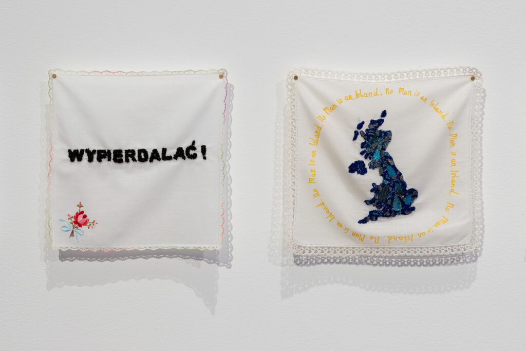 Two white handkerchiefs pinned to the wall. One handkerchief embroidered with the word 'WYPIERDALAĆ' in black thread. The other embroidered with a map of the UK in blue thread, and the phrase 'No Man is an Island' in yellow.