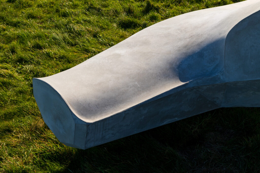 A close-up shot of Ryan Gander's sculpture, showing a grey concrete rectangular shape, with a background of green grass. The concrete resembles a fin or the tail of a whale.