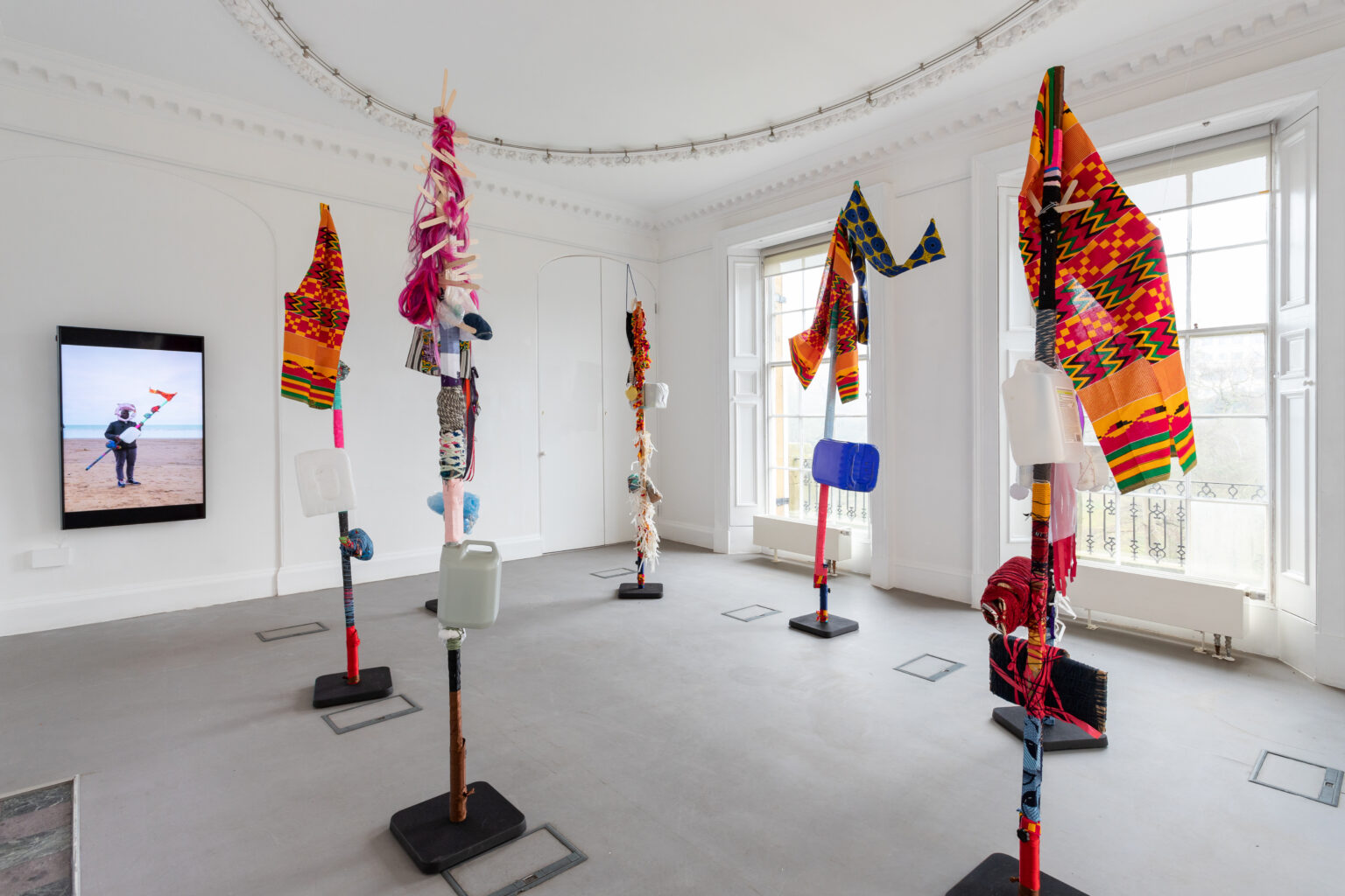A gallery space with six flags on poles in a circle. The flags are made from fabric, ribbons and recycled materials. Mounted on the wall behind, a video shows a figure holding a flag on a beach.