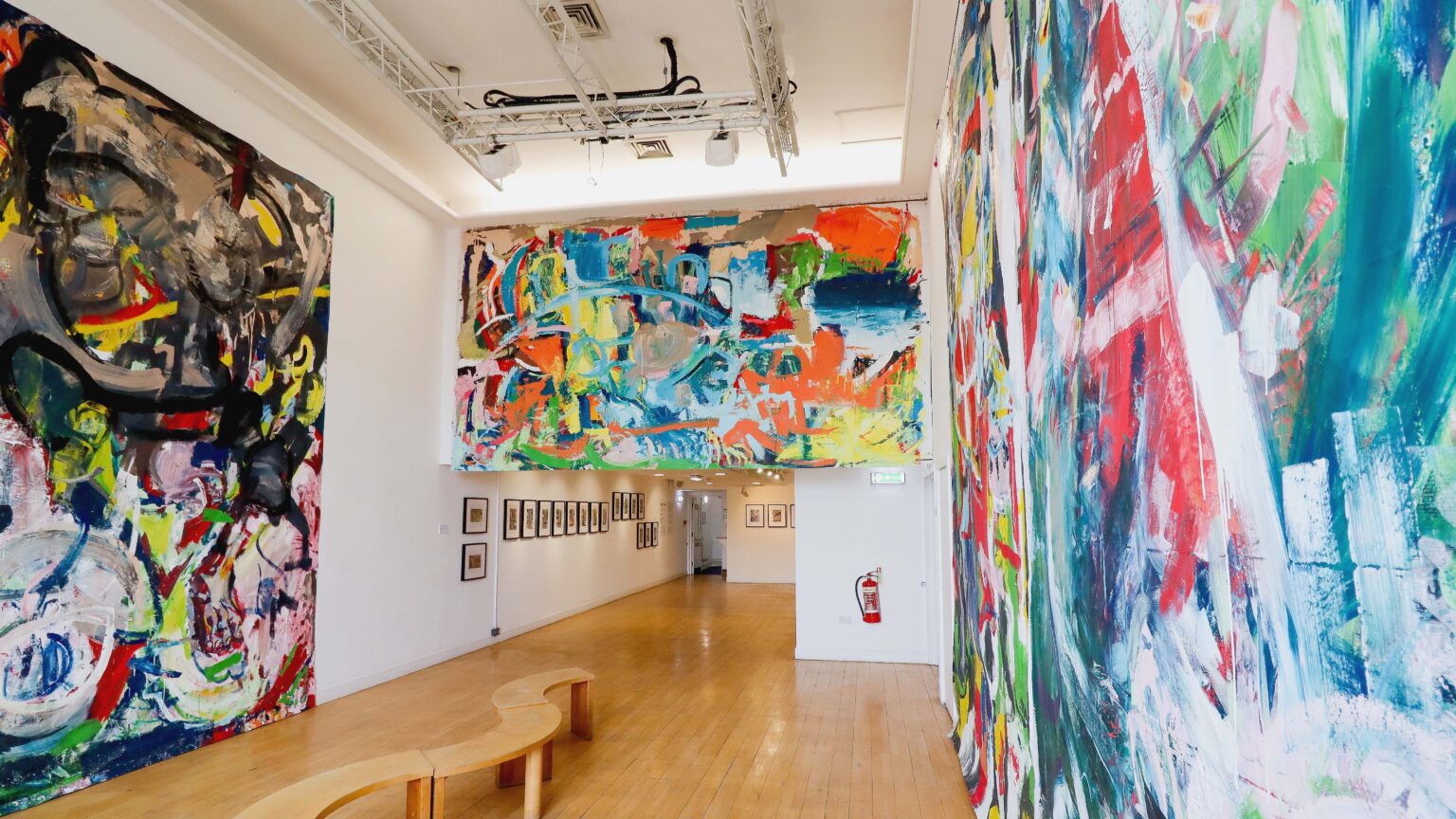 A large, high ceilinged gallery space with a polished pine floor. Large colourful abstract paintings cover most of the wall space. In one section of the gallery are hung small framed photographs.