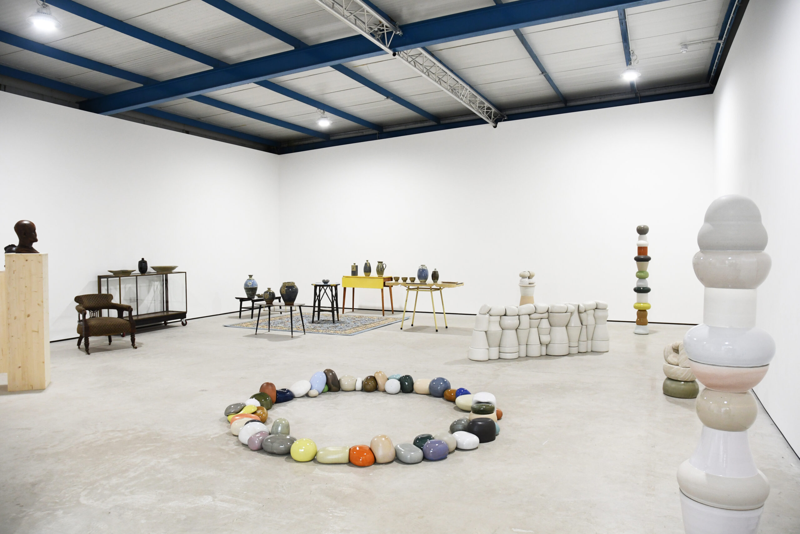 A white cube gallery space is filled with ceramic sculptures. In the middle of the space is what appears to be a circle of multicoloured clay rocks.
