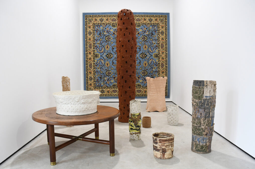 A group of freestanding ceramic forms can be seen in the foreground in front of a rug hung on the wall behind, with one of the sculptures placed on a wooden table.