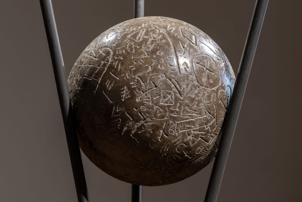 A ball of polished clay, held by three metal prongs. The clay surface is engraved with symbols and marks.