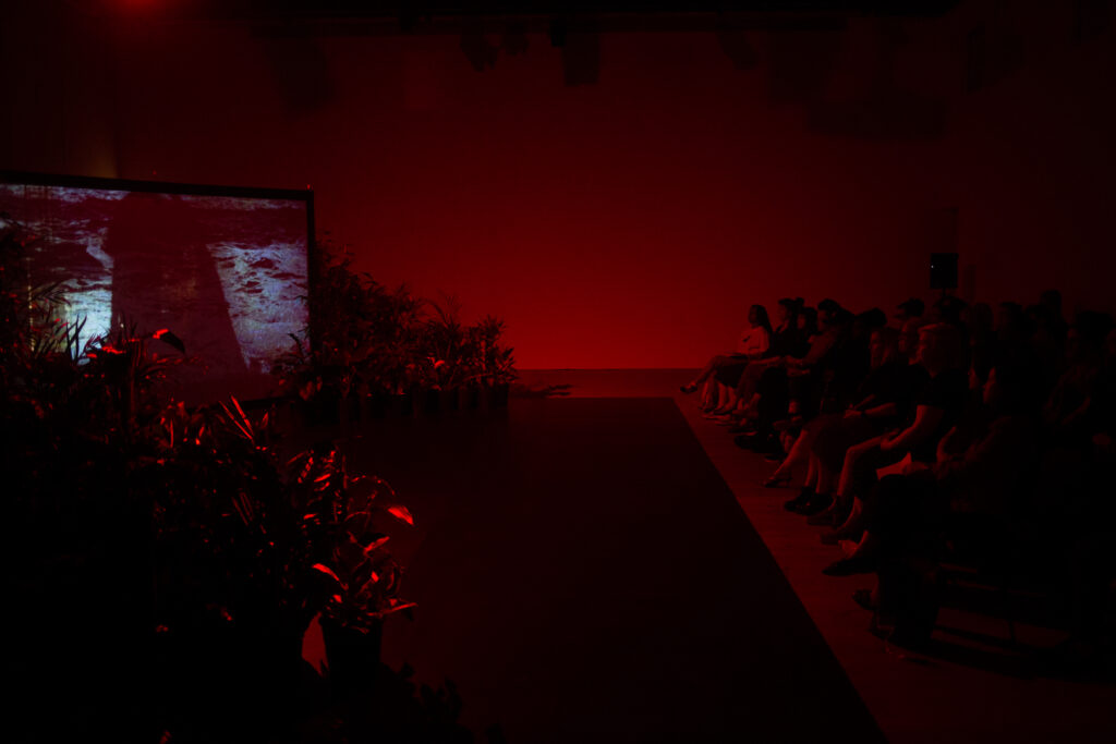 Photograph of the screens and plants set up in the BALTIC space. The shot includes members of the audience watching the screened performance.