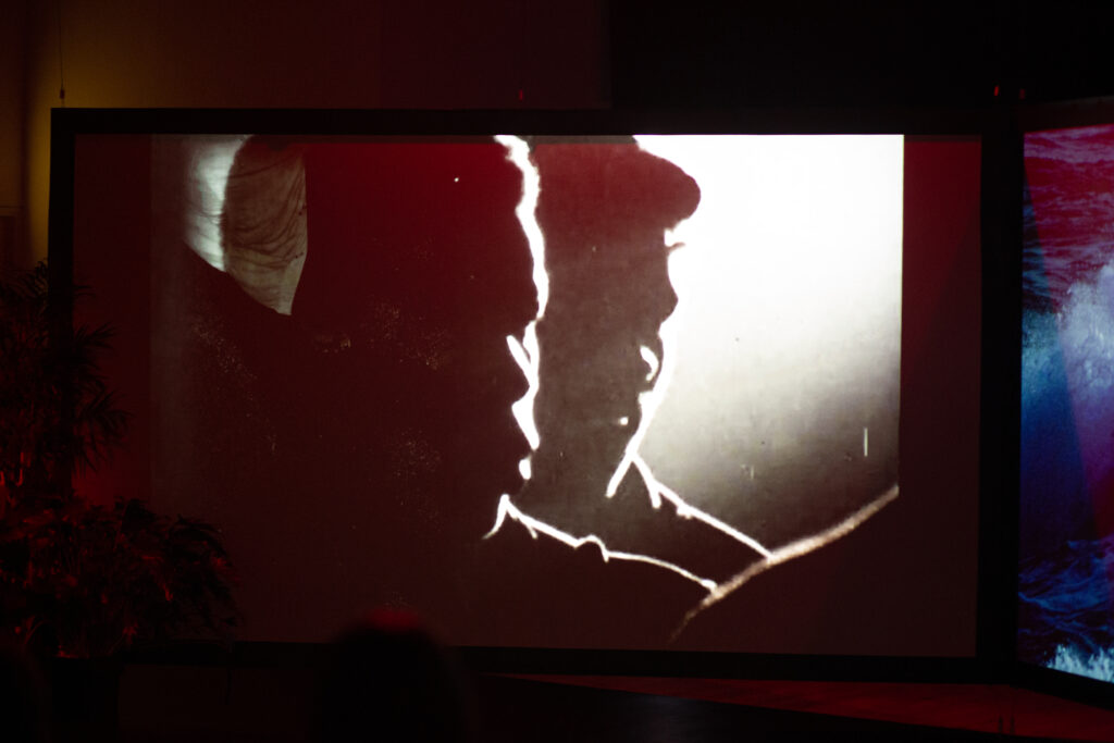 Photograph of the film installation described in the article. on the screen are the semi-silhouetted faces of two men from the Black community. the adjacent screen is just visible and shows sea water crashing.