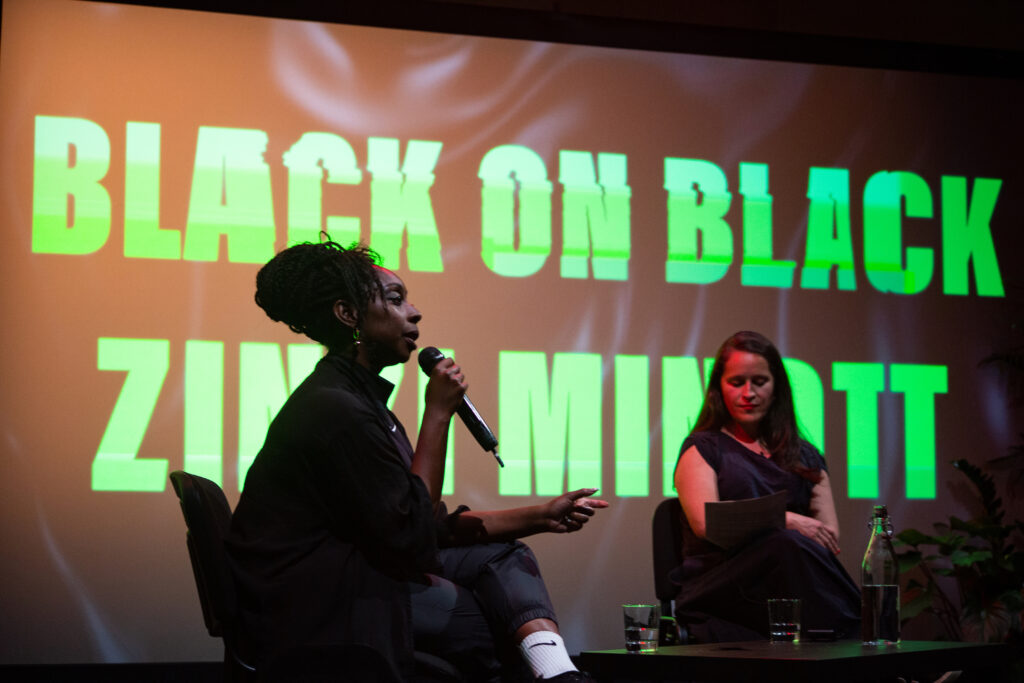 A photograph of the Q and A event described in the article. the artist Zinzi Minnot is speaking into a microphone alongside a curator from the BALTIC. Behind them both is a large screen with the words Black on Black, Zinzi Minnot written in large green letters.