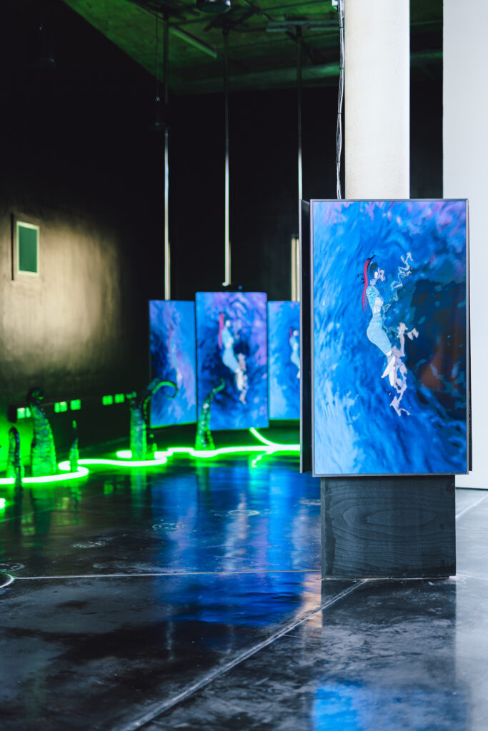 Four monitors in a dark gallery space show an animated person in a pool of water