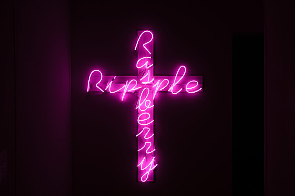 Across made of pink neon strip lights that spells downward 'raspberry' and across 'ripple' against a black background