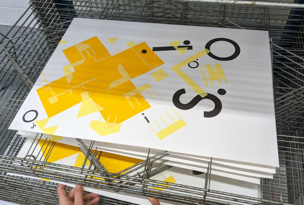 A stack of prints on a drying rack in a printmaking studio, with yellow and black designs combining abstract shapes and letters.
