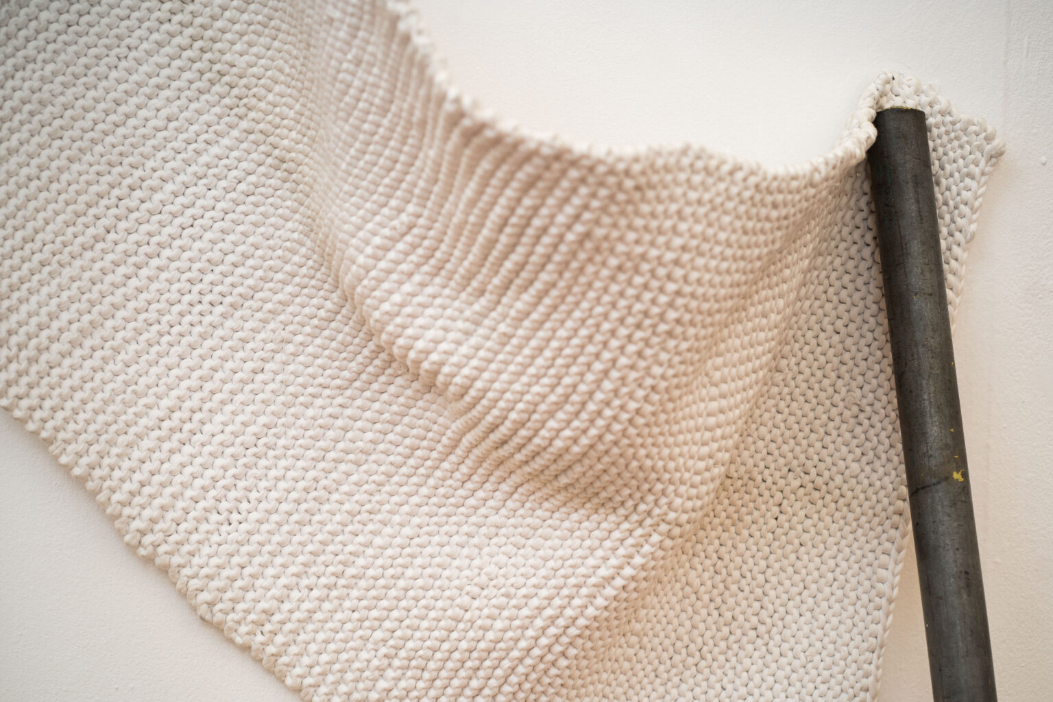 A draped piece of white knitted fabric is pinned against a white wall by a silver scaffold pole.