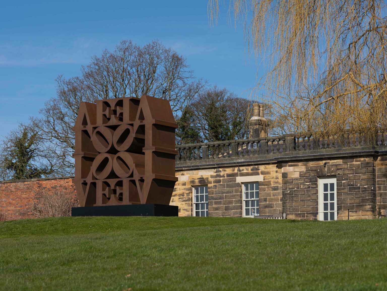 A corten steel sculpture made up of the letters 'LOVE' repeated and stacked in a square format. The sculpture is set on a grassy mound with a stone building behind.