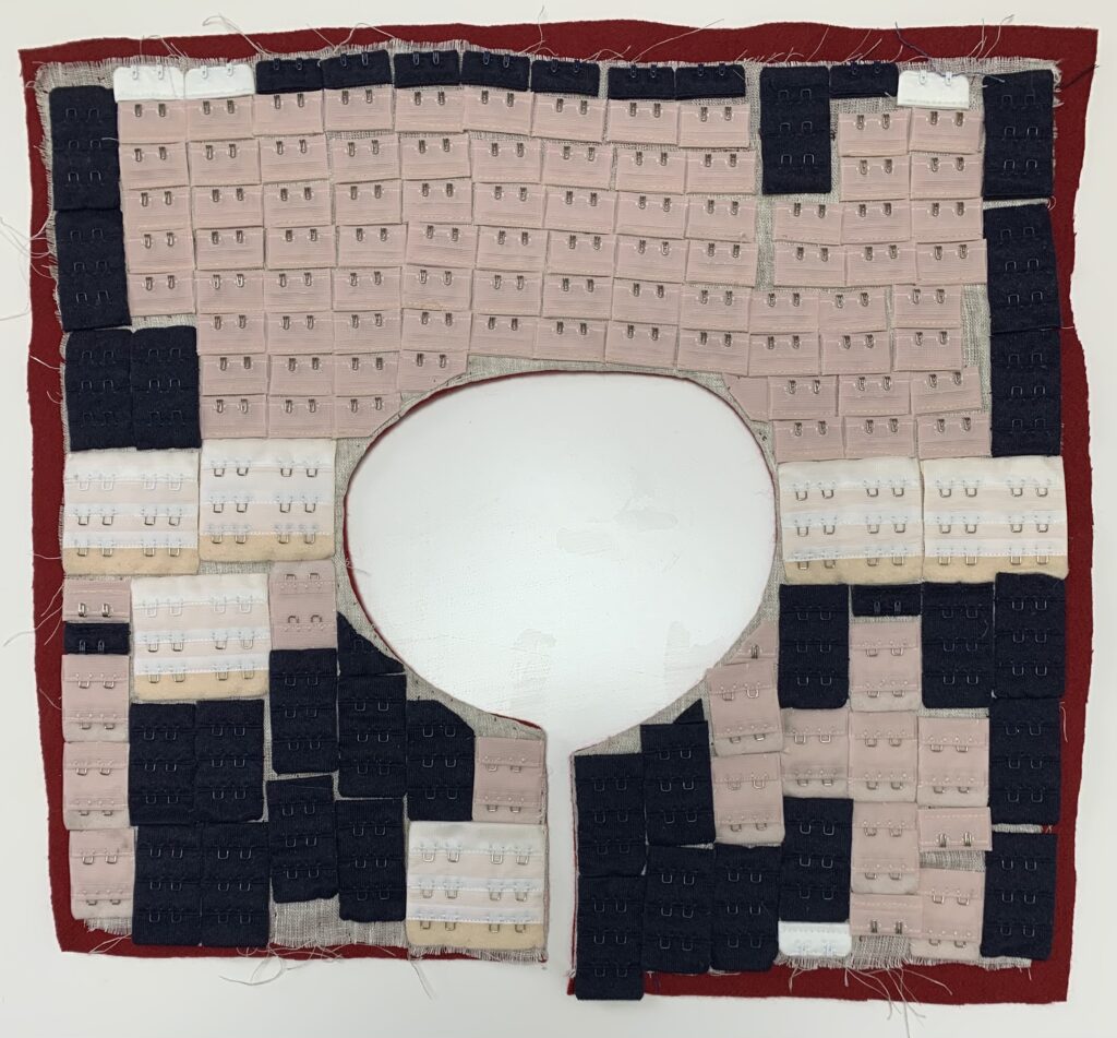 A square of red felt with hook-and-eye attachments sewn on in a pattern of light pink, white and black, with a head-size circle cut out of the middle.