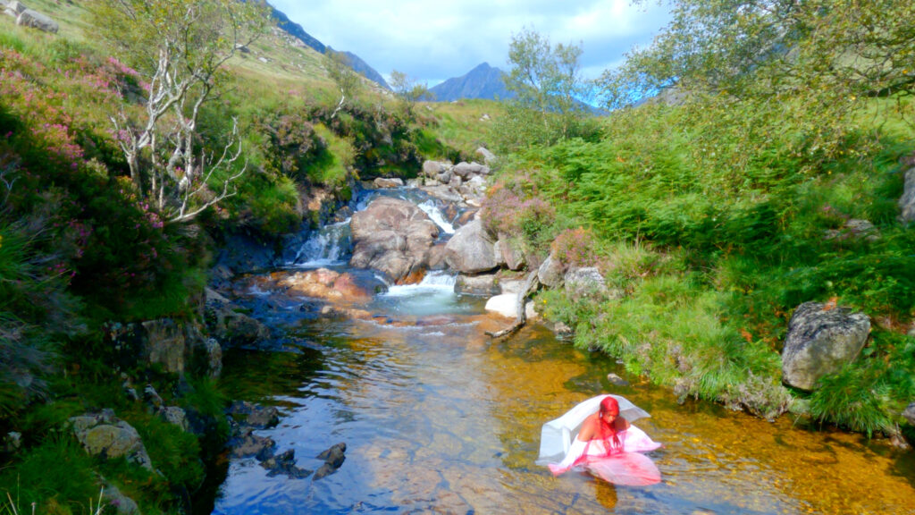 A person dressed in white kneels in a river. They have bright red pigment on their hair, dripping down onto their white costume and into the water. The surrounding landscape includes rocks, heather and silver birch, with mountains in the background.