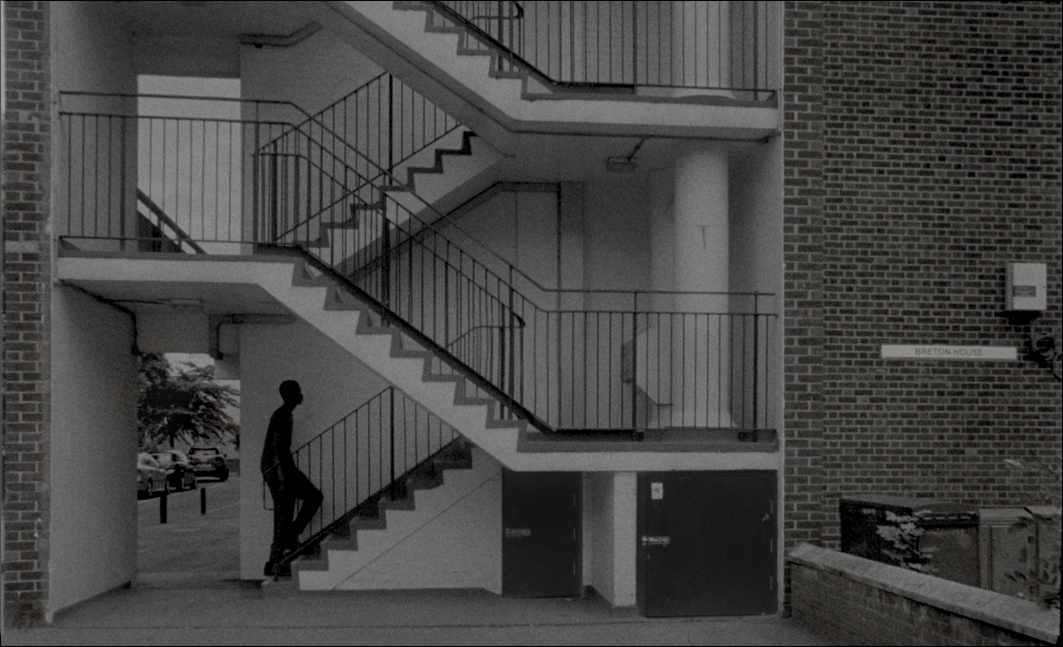 A black and white film still, in which a figure is starting to ascend a staircase in a south London tower block.
