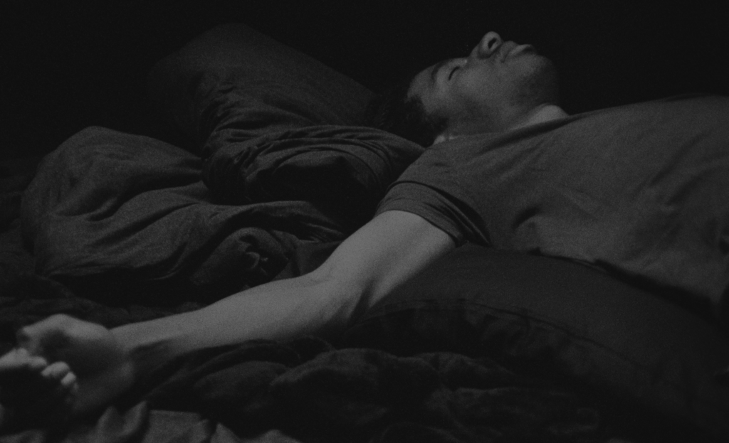 A black and white film still showing a person sleeping. Their eyes are closed, their face is peaceful, their arm stretches out across the frame.