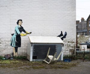 A discarded fridge freezer against a brick wall with a painted woman beside it and man's legs sticking out