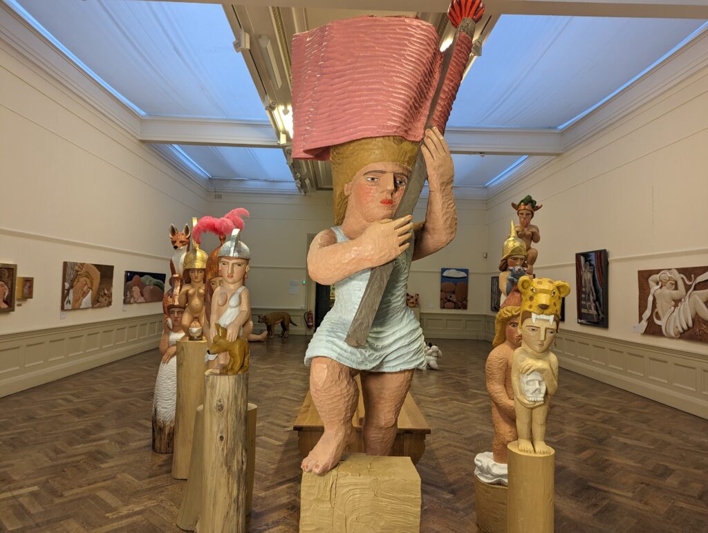 A wood carved sculpture of a white woman carrying a flag on a plinth with other works behind