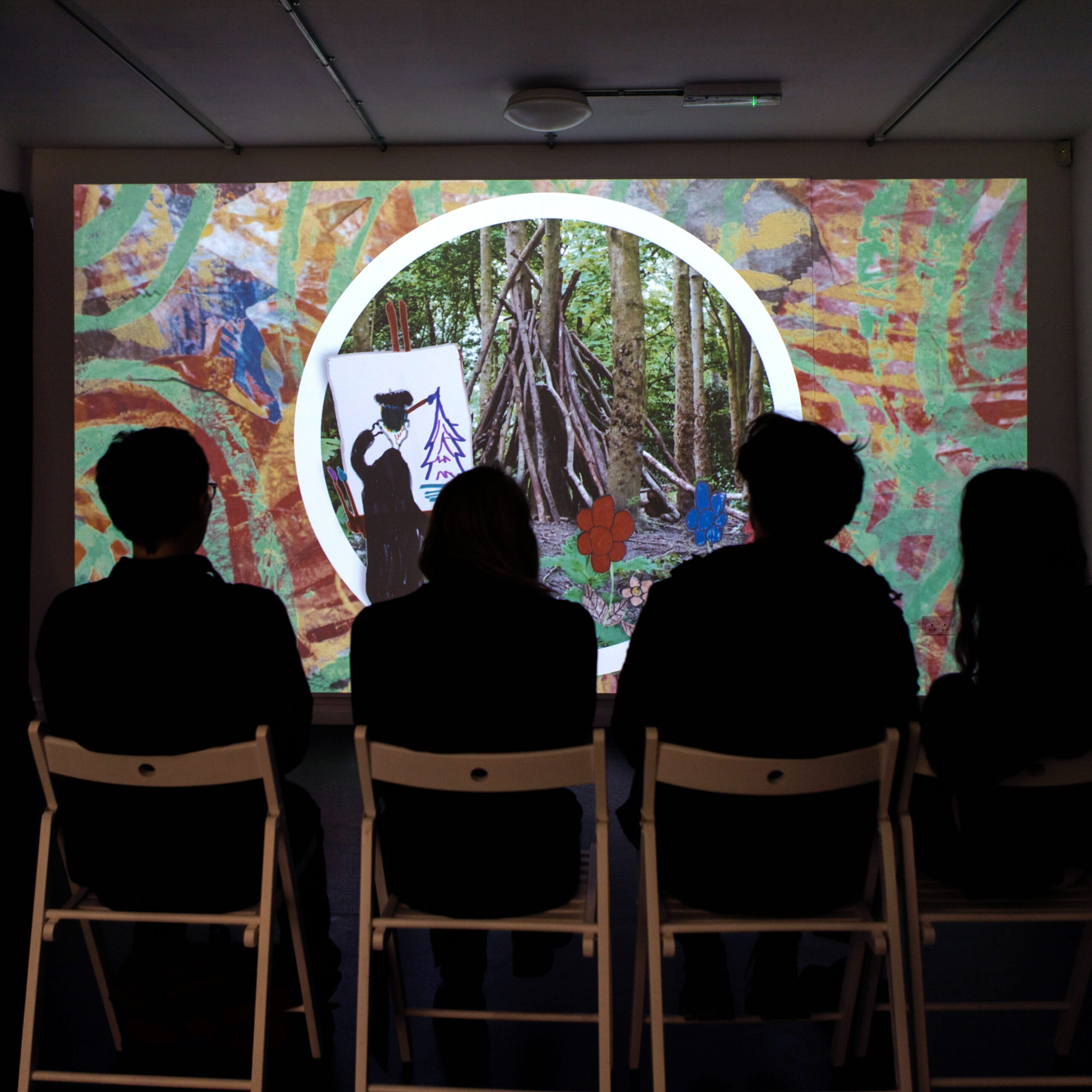 Four people are seated in front of a large projection screen at Brighton CCA Dorset Place; they are silhouetted in a darkened room. On the screen is a still image from 632700, showing an red and green abstract pattern around the edge, a central white circular band with a photograph of a den made from sticks in the woods inside the circle, a drawing of red and blue flowers in the bottom right, and a drawing of a person painting a landscape scene on canvas in the bottom left.