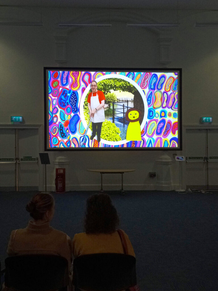 Installation shot of 632700 in the Henry Moore Lecture Theatre at Leeds Art Gallery. The room is darkened and the backs of two people watching a large projection screen can be seen in the bottom left corner. On the screen is a still from 632700 showing an abstract background of brightly coloured blobby shapes, reminiscent of amoebas. There is a central circular band with a photograph of a river, a railing, and lots of leafy plants inside. On the bottom right of the image is a drawing of a lion with a friendly face in coloured felt pen. On the left of the image is a photograph of a white man in a red t-shirt and an apron, holding a golf club made from tin foil about to hit a pink ball.