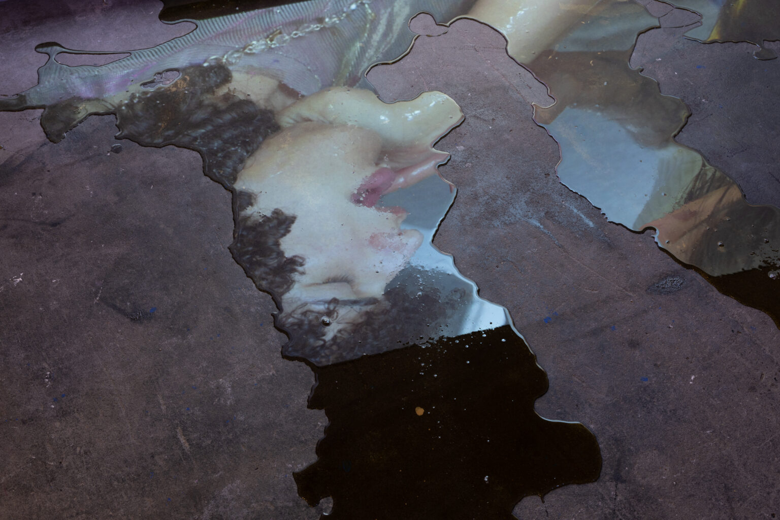A fragmented still from a film is reflected in dark liquid seeping on the gallery floor. The reflected image is upside down, but it shows a person with their eyes shut and mouth open.