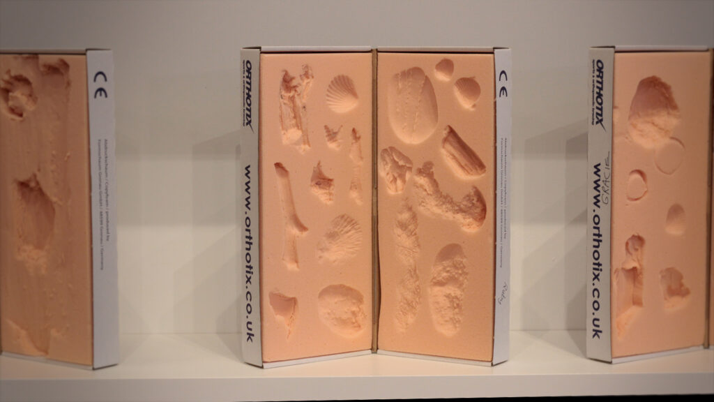 Three blocks of pale pink foam on a white shelf that show the impressions of various coastal objects including shells, sticks and pebbles.