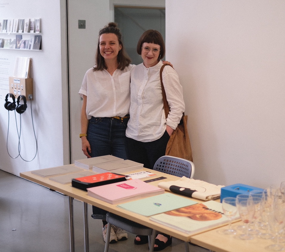 Photograph of two smiling women in white shirts standing behind a table with colourful publications and wine glasses, in a gallery foyer.
