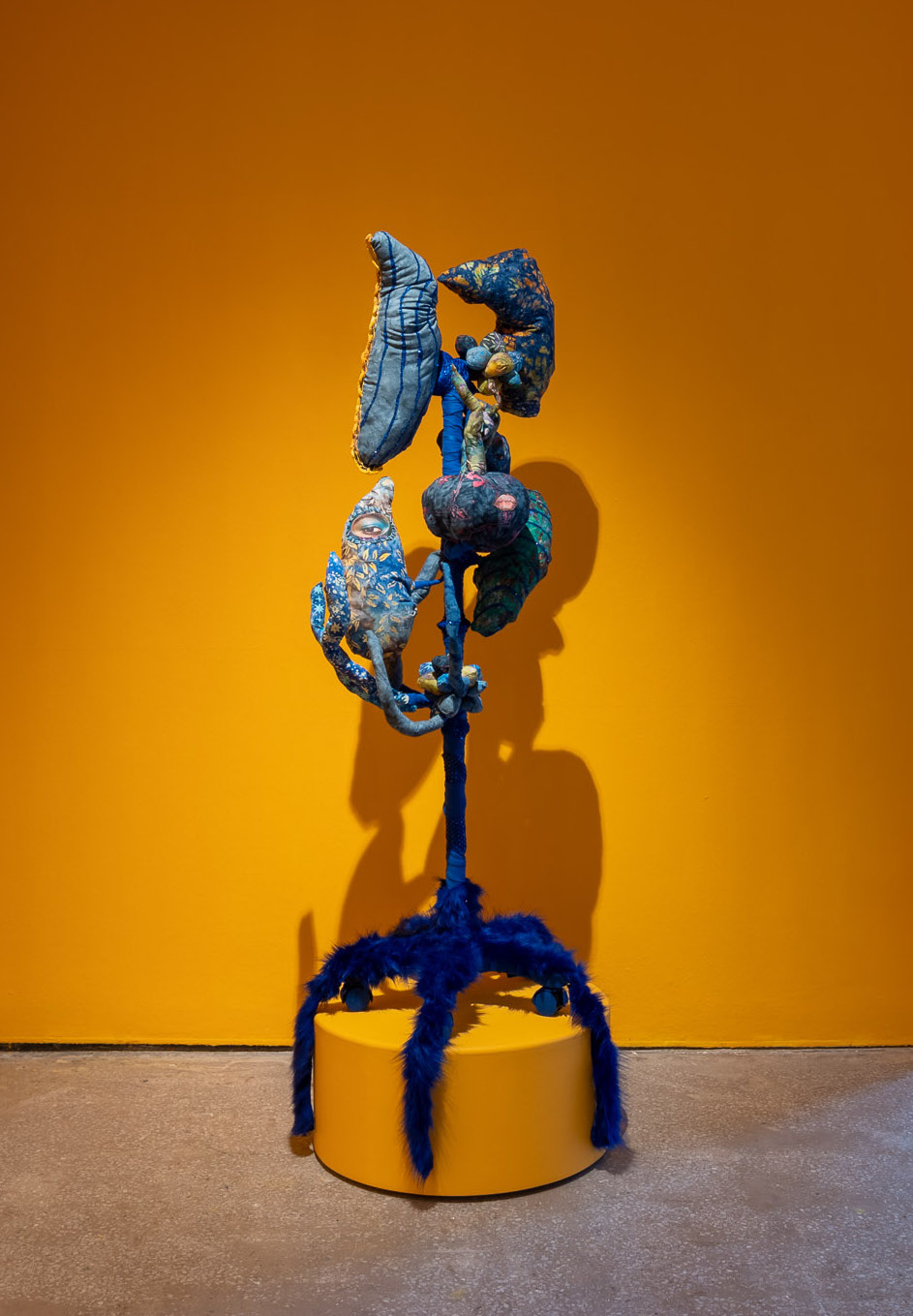 An abstract-figurative sculpture perches on a deep yellow circular plinth in a yellow painted gallery corner.