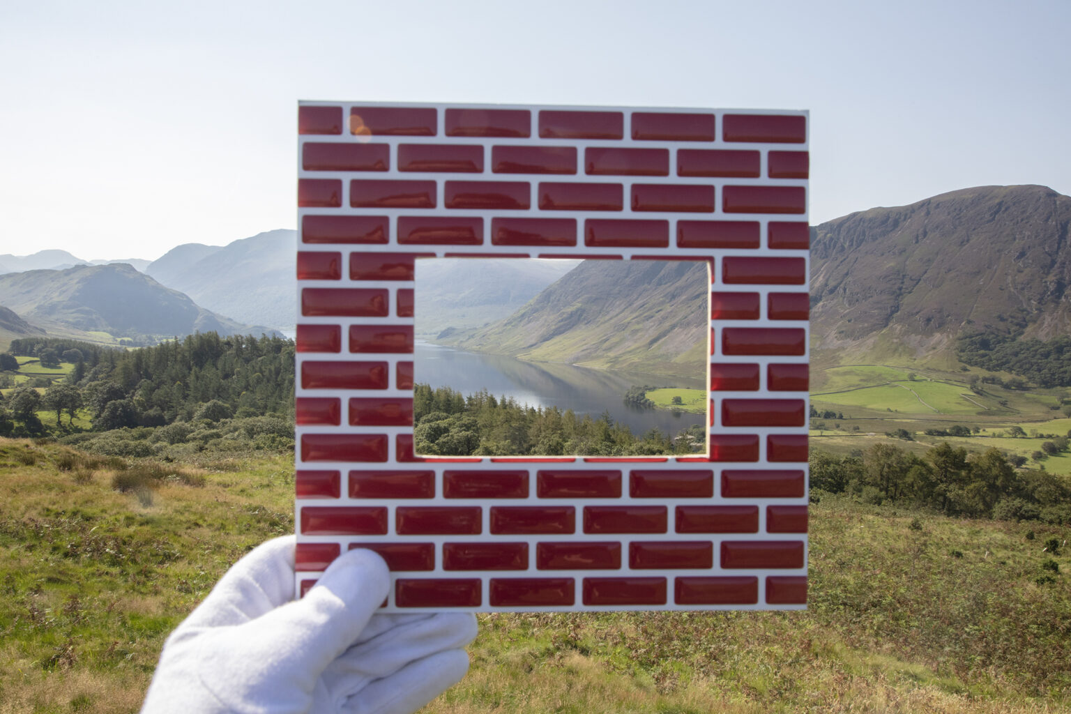 A white gloved hand holds a viewfinder, made to resemble a brick wall, up against a majestic landscape.